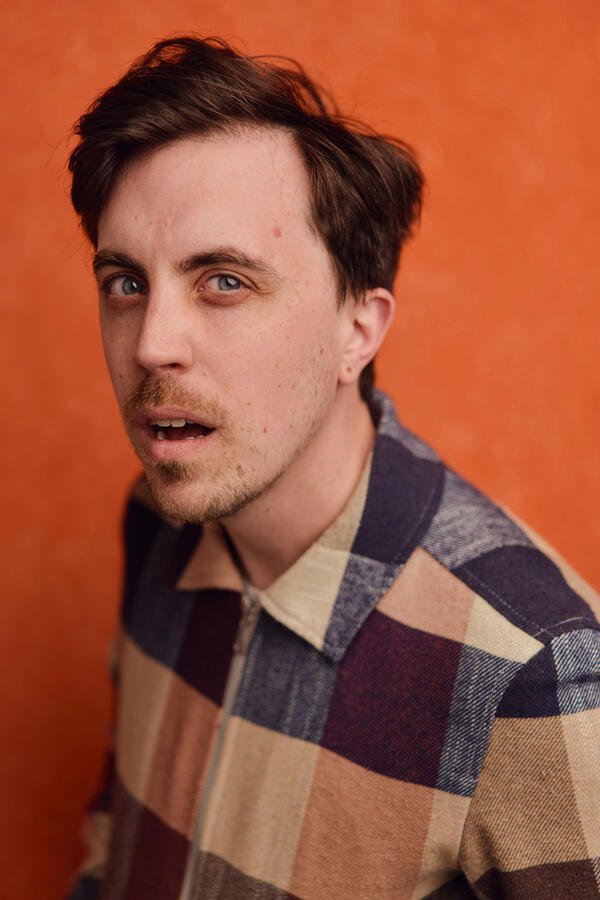 Headshot by Olivia Spencer Felix Trench in a multicoloured jacket on an orange background.